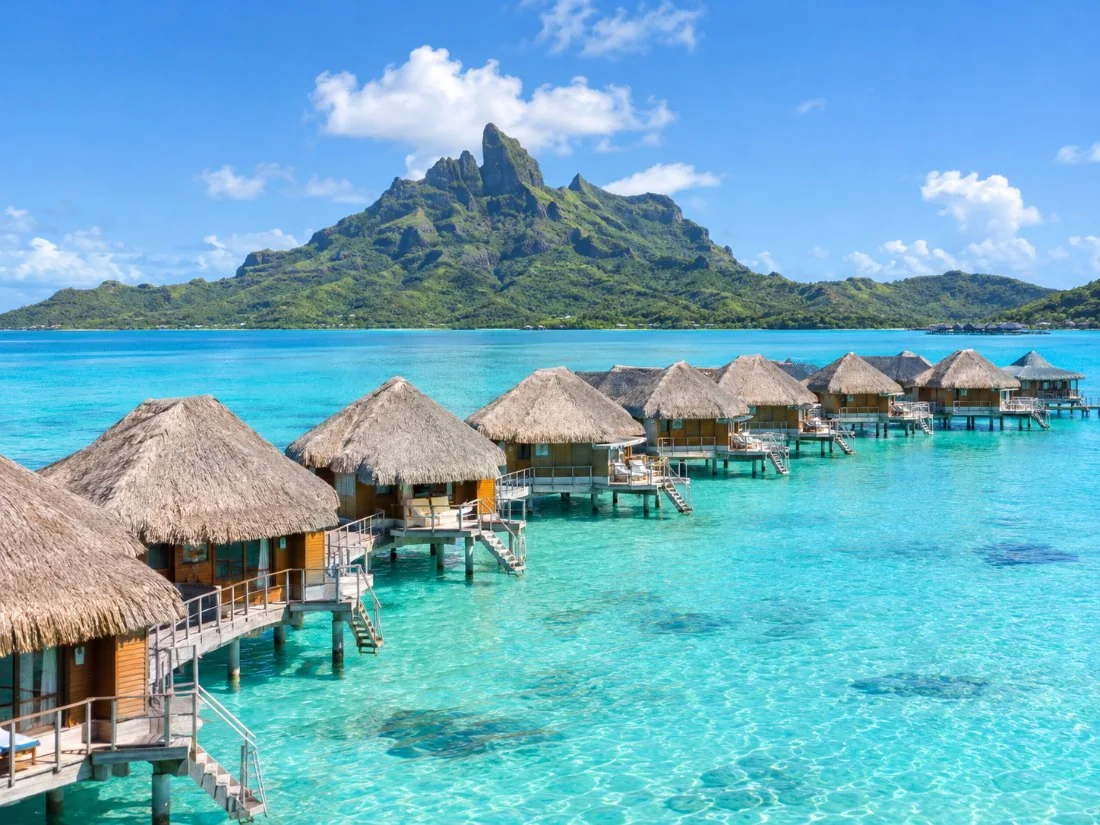 A row of overwater bungalows on stilts over pale turquoise water inside the Bora Bora lagoon