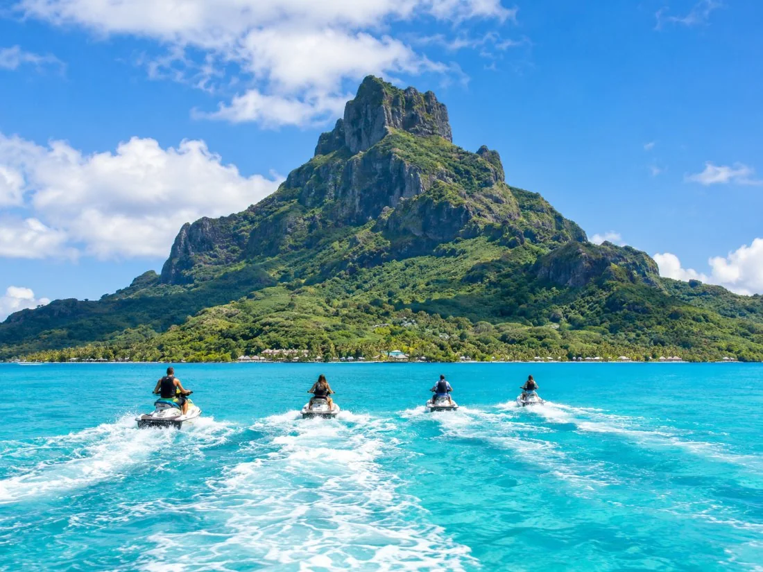 Jet skis riding in a line across the turquoise lagoon with the dark green peak of Mount Otemanu behind
